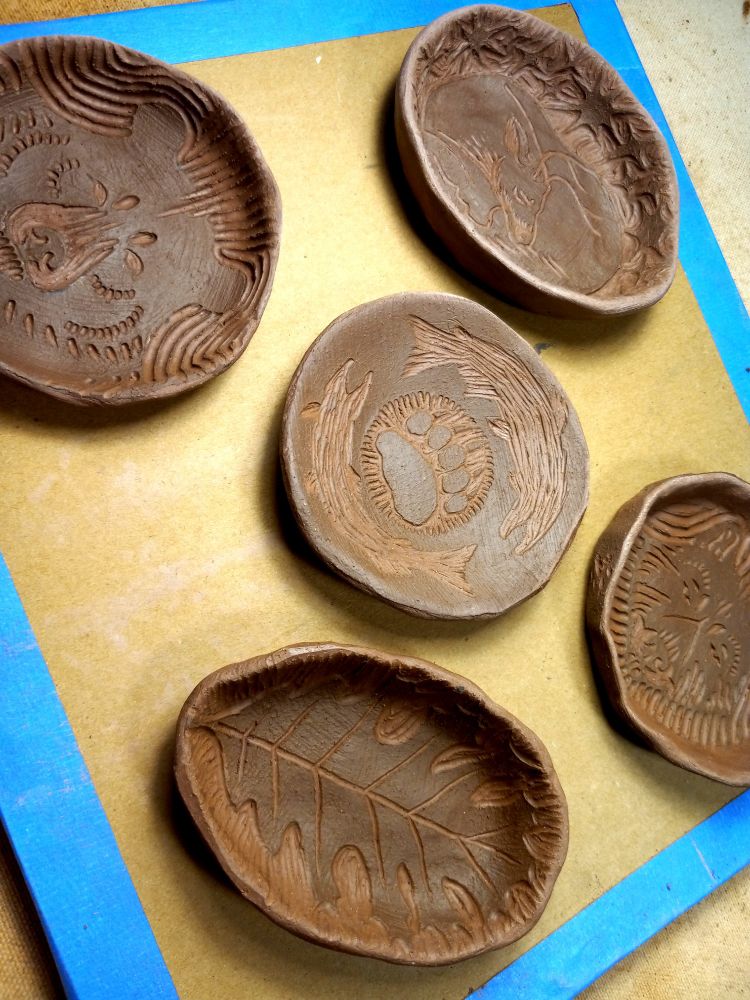 Photo of five different small clay dishes with carvings of various animals and a leaf. The top layer of clay is slightly darker in tone than the main clay body which is a pale red.
