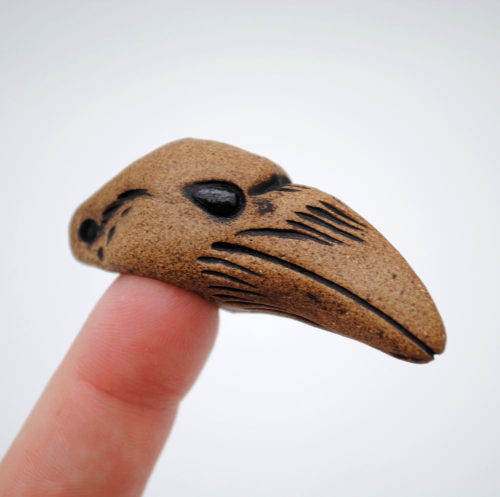 photo with white background of a fired clay pendant perched on a fingertip, the shape of a raven's head