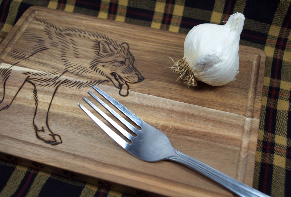 photo showing a small wooden cutting board with a woodburned image of a walking wolf on the left side. the board also has a head of garlic and a fork sitting on it. the board is sitting on top of a black yellow and red plaid fabric