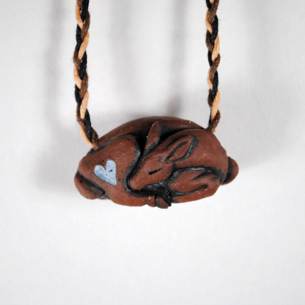 photo with white background of a red clay pendant hanging on a woven cord, the pendant is in the shape of a sleeping doe with a light blue heart on her thigh