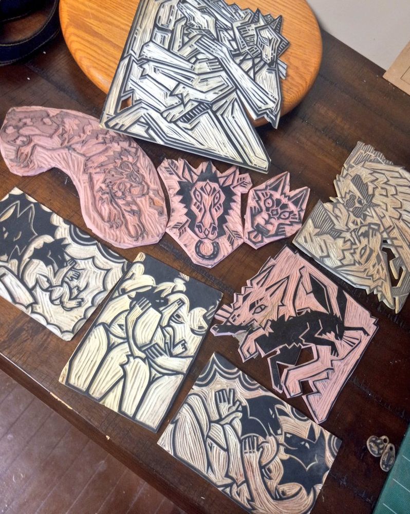 Photo of a number of linoleum carved blocks for making prints sitting on a tabletop, with various designs like jumping tiger, horse and canine heads, wolf headed trans figures, a wolf and rabbit, etc. 