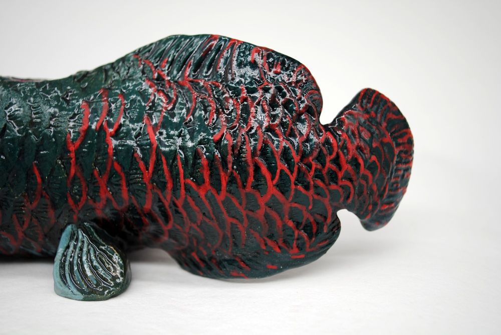 close up cropped photo of a ceramic pipe with a white background, the pipe is in the shape of a arapaima fish. This photo shows the details and red scales of the tail.