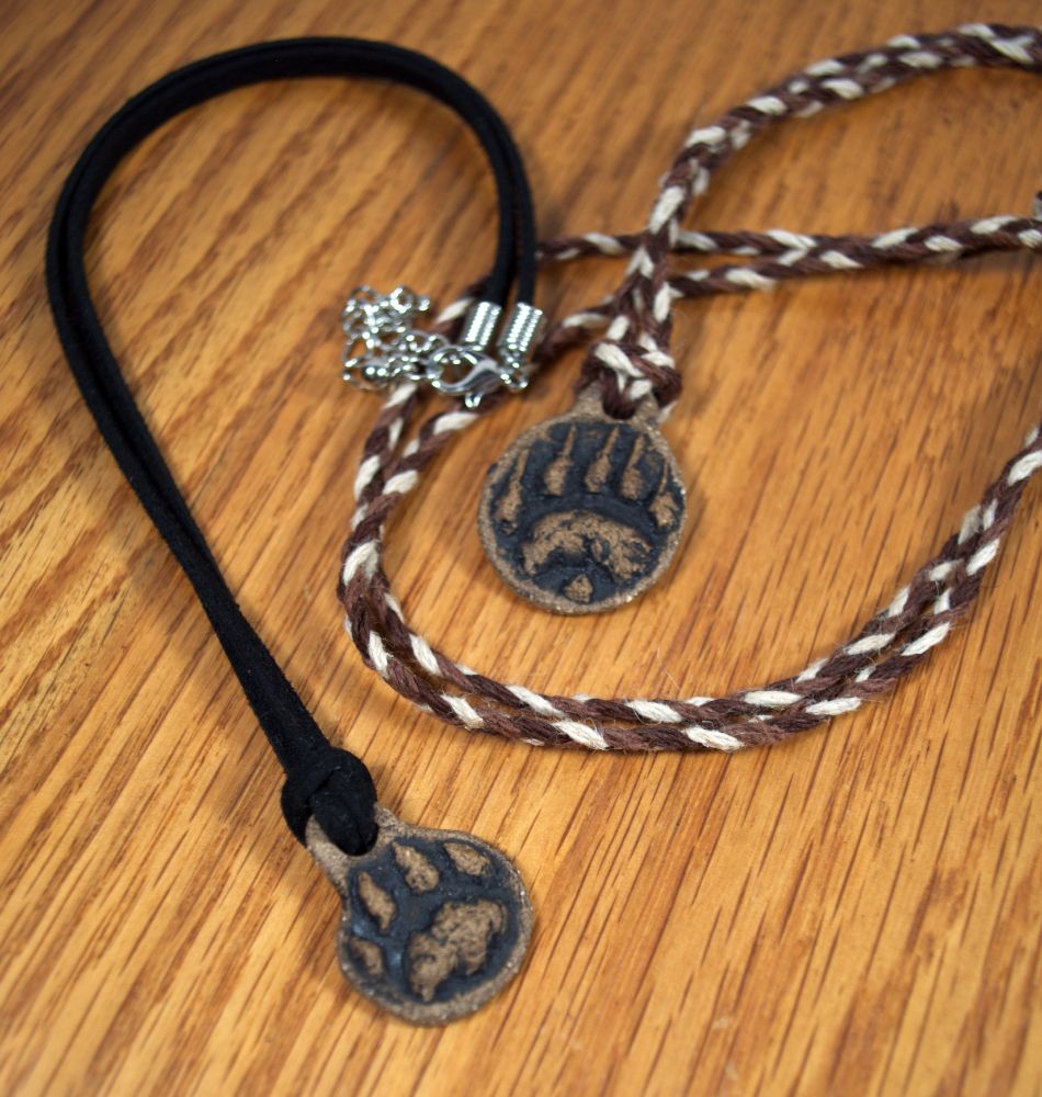 photo showing two paw print pendant necklaces, one on a black suede leather cord with a big cat paw, and the other with a braided brown/cream cotton cord with a bear paw. they are sitting on a wooden surface.