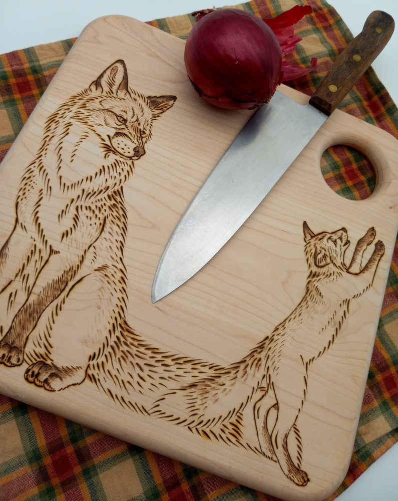 A photo with white background of a wooden cutting board sitting on a autumn colored plaid cloth with a red onion and knife sitting on top of the board. The board has the image of a adult red fox watching her kit play