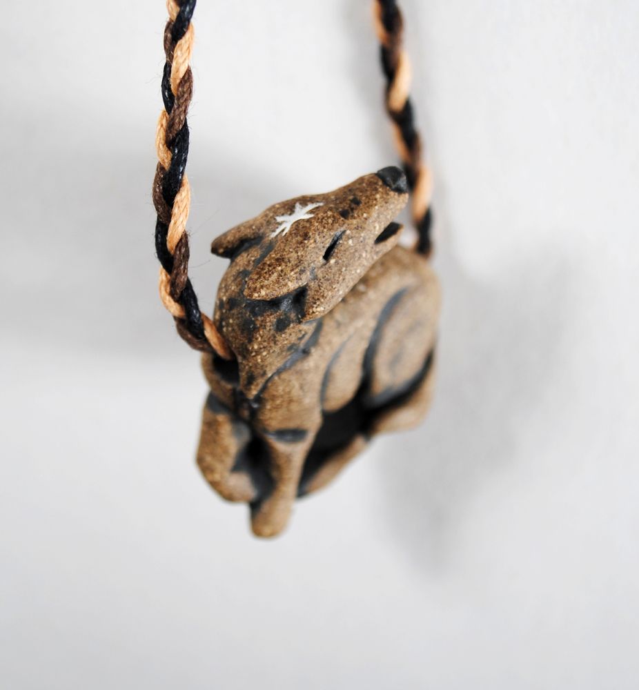 photo with a white background of a warm grey clay pendant in the shape of a standing howling wolf on a multicolor cotton cord, focusing on a white star marking on the forehead