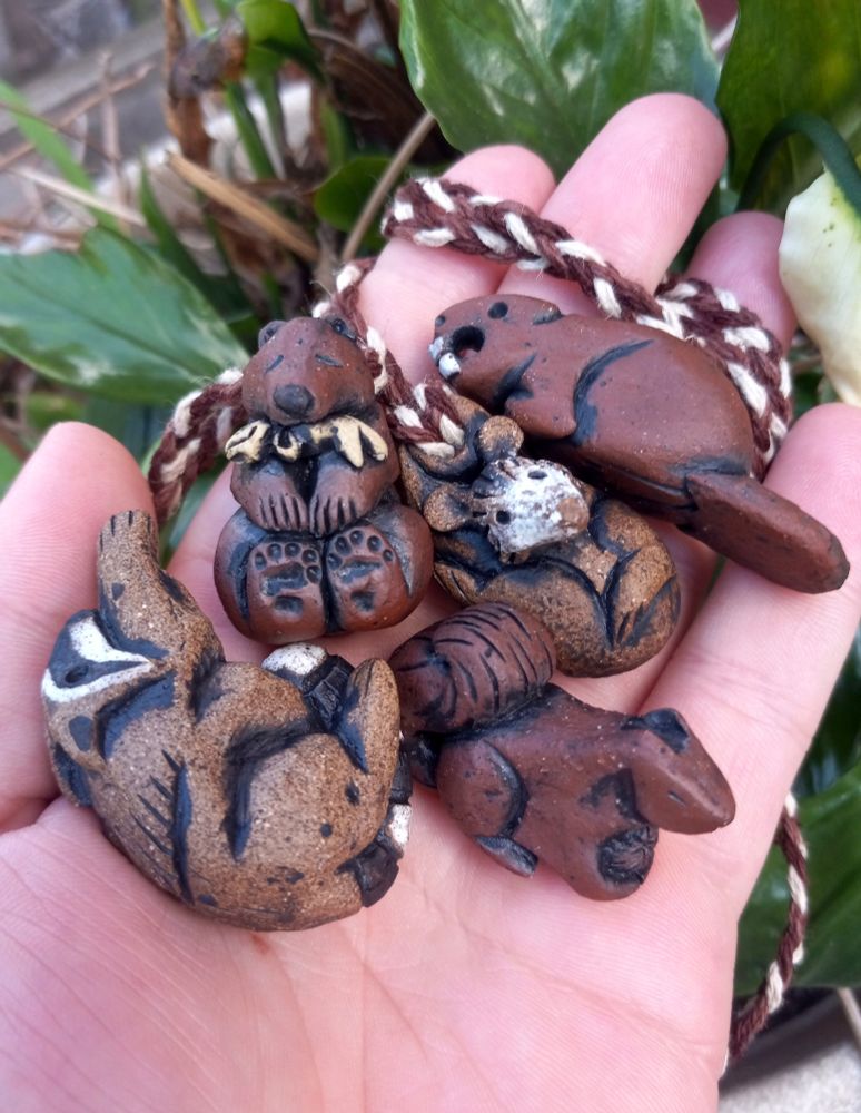 Photo showing a hand in front of green plants, in the palm of the hand (left to right) is little clay pendants in animal shapes, a raccoon, a bear with a fish in its mouth, a squirrel holding an acorn, a hanging opossum, and a beaver 