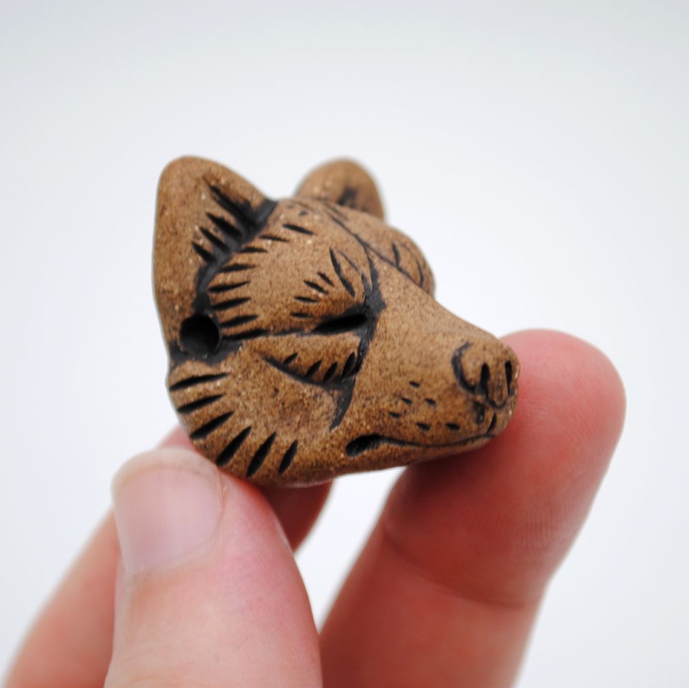 photo with white background of a fired clay pendant perched in three fingers, the shape of a wolf head