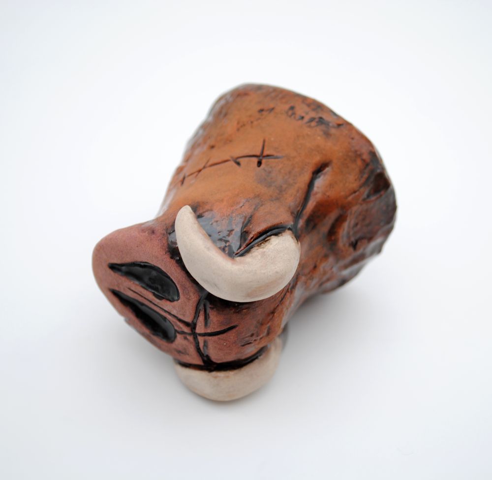 photo with white background of a ceramic shot glass in the shape of a pig/boar snout sitting at an angle to show the bottom details. the snout is covered in scars and has glaze in shades of sandy brown to pink, with white curved tusks