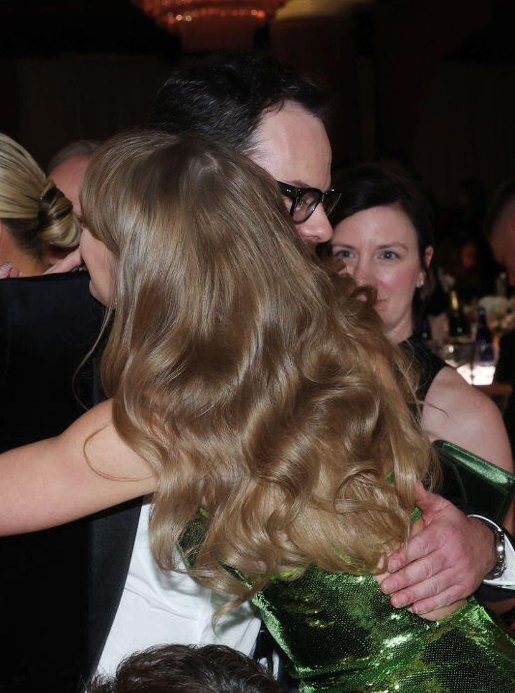 taylor swift at the 2024 golden globes - this picture is a view of her back. you can see part of her green sparkly dress but her wavy long blonde hair is covering most of it. she's hugging a man and you can tell she's smiling.