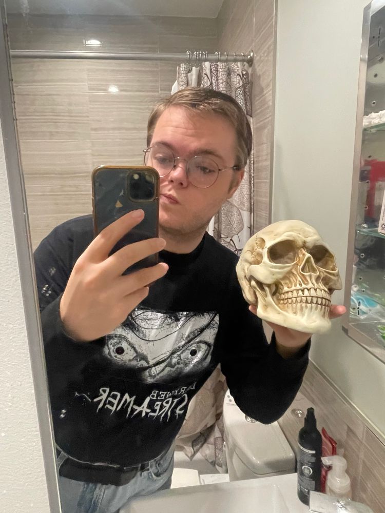 caucasian person with blonde hair, round glasses and a spooky t shirt taking a selfie with a skull.  