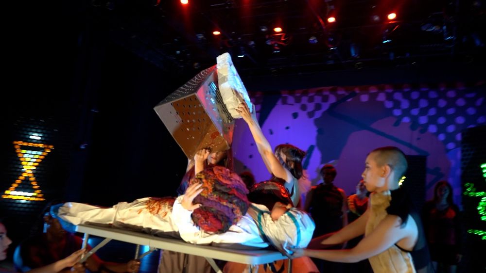 A woman uses a giant fake cheese grater to grate fake cheese over another woman at a dance performance.