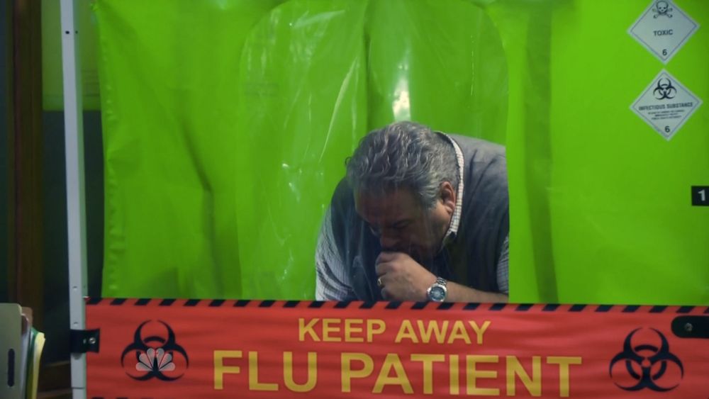 Jerry Gergich, a character on the TV show Parks and Recreation, coughs in a bright green quarantine tent. On the front is a red sign with large yellow text that reads: KEEP AWAY, FLU PATIENT. 