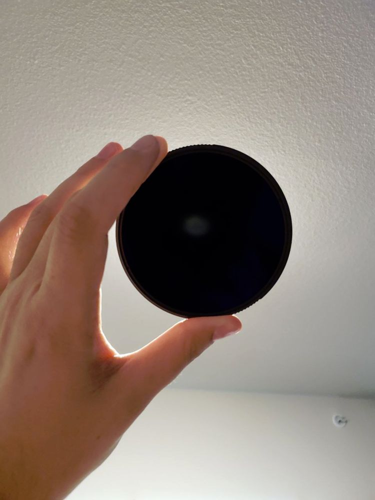 Another photo of my hand holding up a solar filter for a camera lens.  This time I am holding it in front of an overhead light fixture in my apartment.  The light from the fixture is just barely visible as a dim white blur through the filter.
