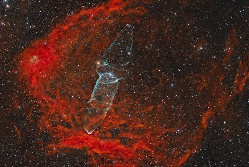 A nebula featuring a blue squidlike structure in the center, which is surrounded by a red semicircular gas cloud. The surrounding gas is faint in the center and brightens towards the outer edge. 