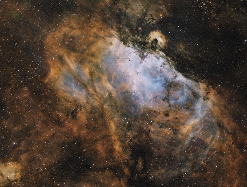 A warm gold colored nebula with a bluish white core region. The core contains a famously photographed region commonly referred to as the pillars of creation. 