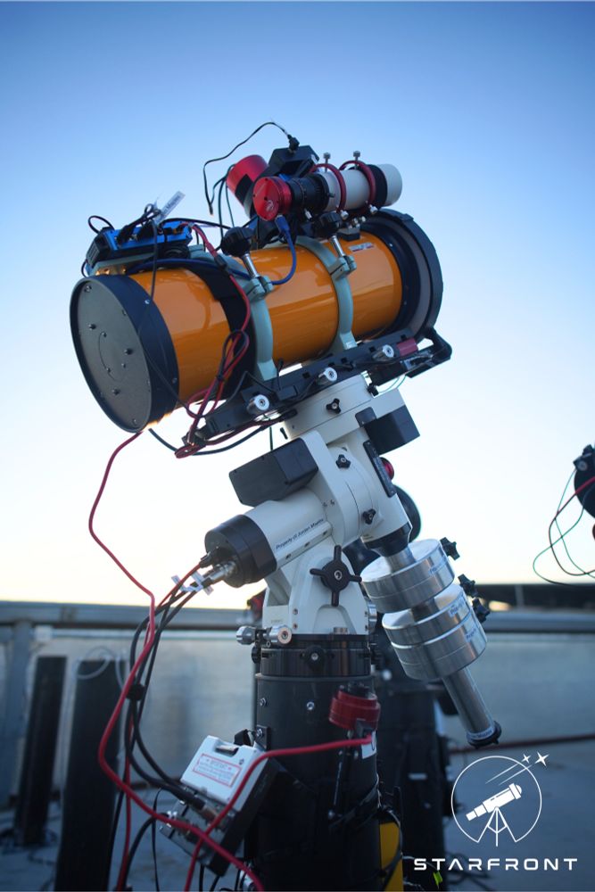 A yellow reflector telescope attached to a white mount with stainless steel counterweights sitting atop of a black steel pier. The scope is inside a building with a roll-off roof which is open, revealing a clear blue sky in the background. There are other, unoccupied piers in the background.