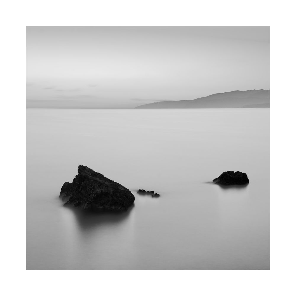 A black and white long exposure of some boulders in the Mediterranean with a sliver of land in the distance. © Damian Ward 2025. 