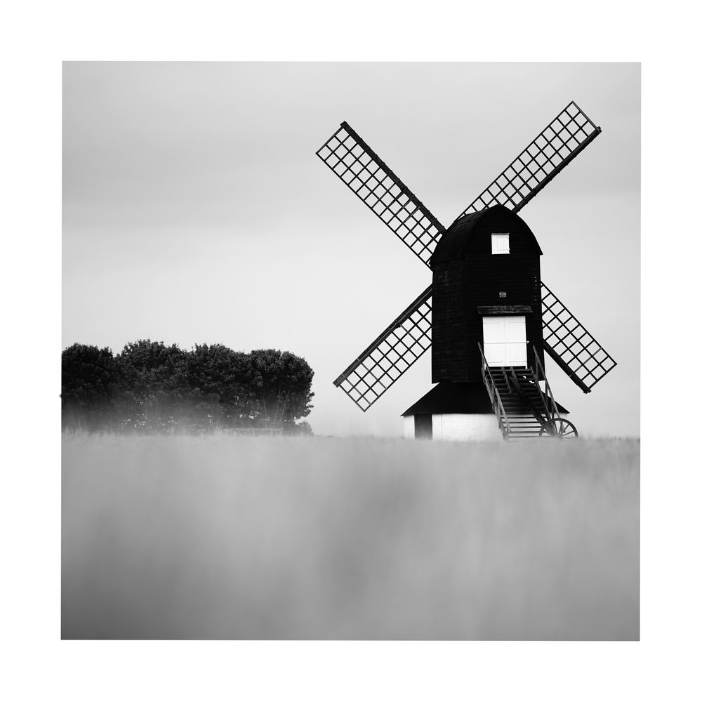 A black and white image of the windmill at Pitstone. The image is low down with a blurred crop at the bottom and a dark line of trees on the left. © Damian Ward 2025. 