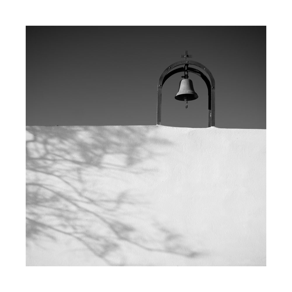 A black and white image of a chapel bell and its bright white wall. The wall has a shadow of a nearby tree taking up the lower left third. © Damian Ward 2025. 