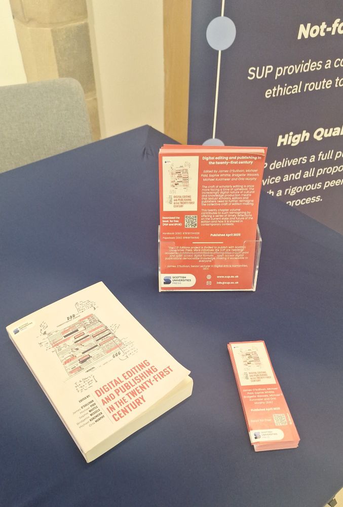 A photograph of display table with a dark blue tablecloth, a copy of the book Digital editing and publishing in the twenty-first century and some flyers and bookmarks