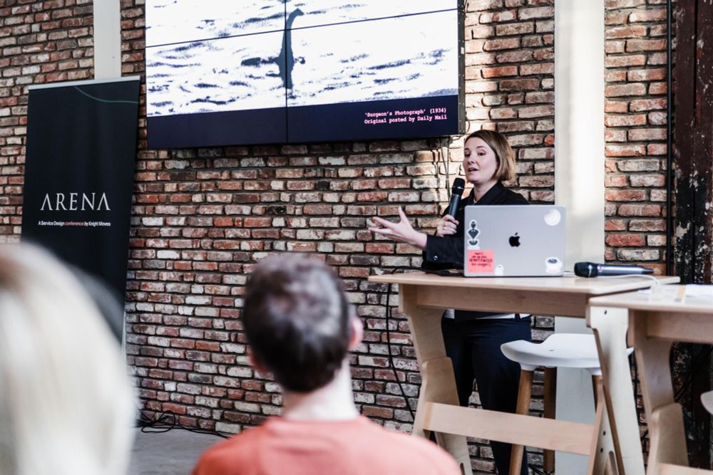 A person in navy blue giving a talk in front of a screen and brick wall 