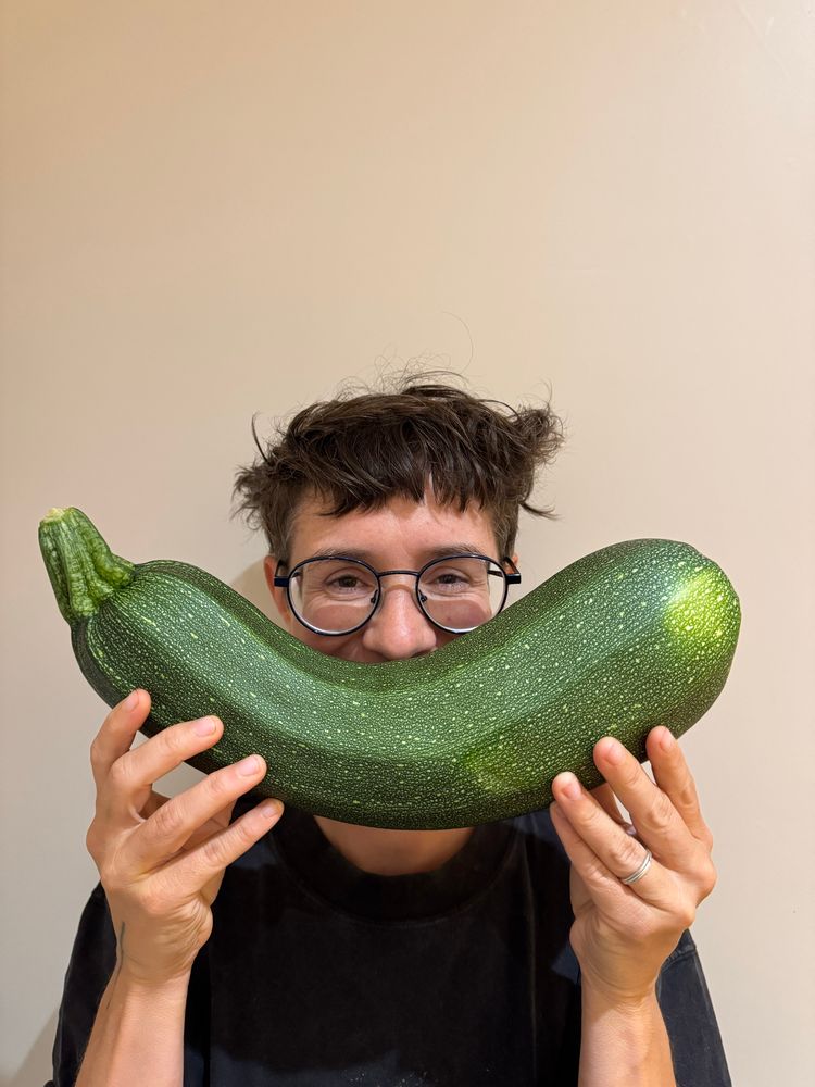 A person with circular glasses holding up a green courgette that is massive as a fake smile 