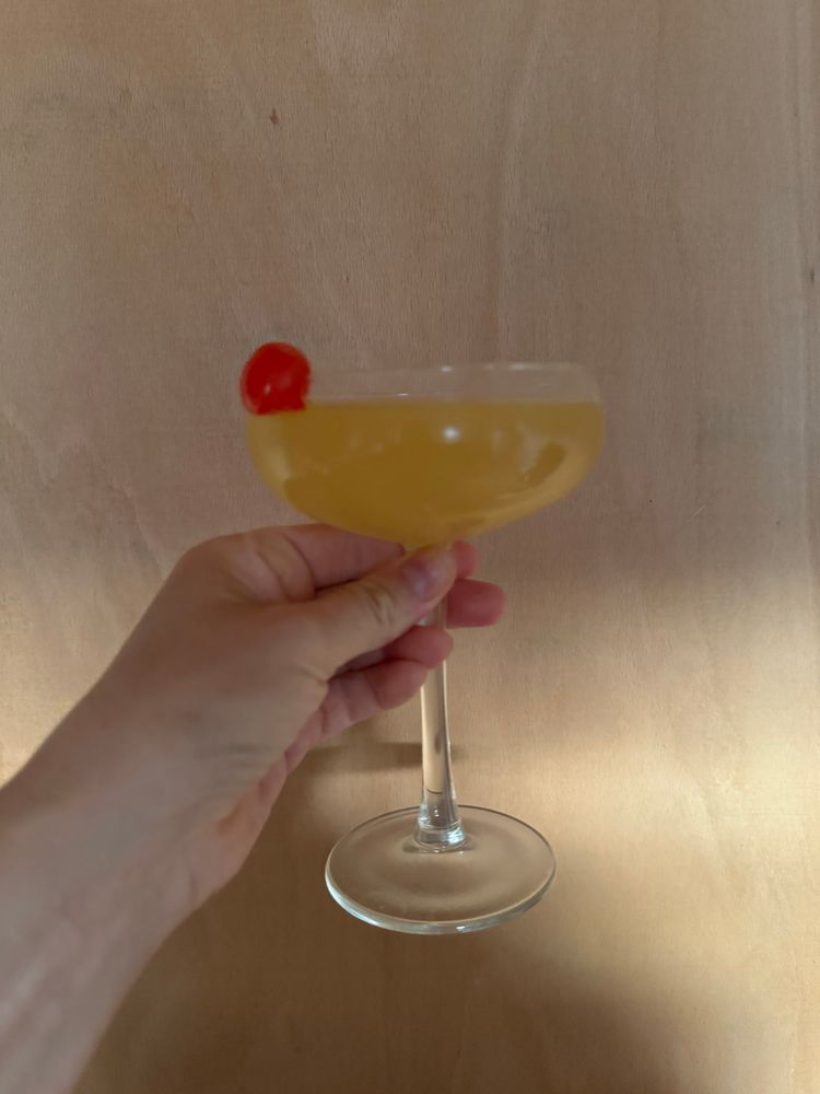 A glass with yellow liquid in it and cherry in the side being held up by a hand 
