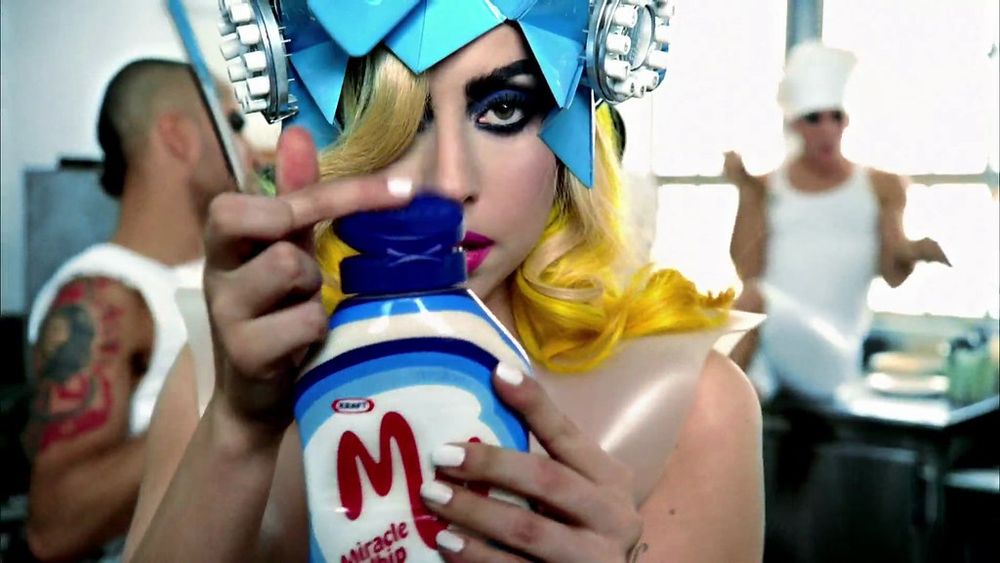 A screenshot of a lady gaga music video where she is holding a modern container of Miracle Whip, and a knife