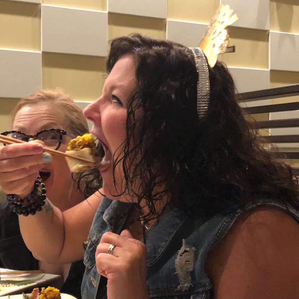 Restaurant Scene: Woman, mouth wide open, about to place huge roll in her mouth while an astonished friend looks on. 
