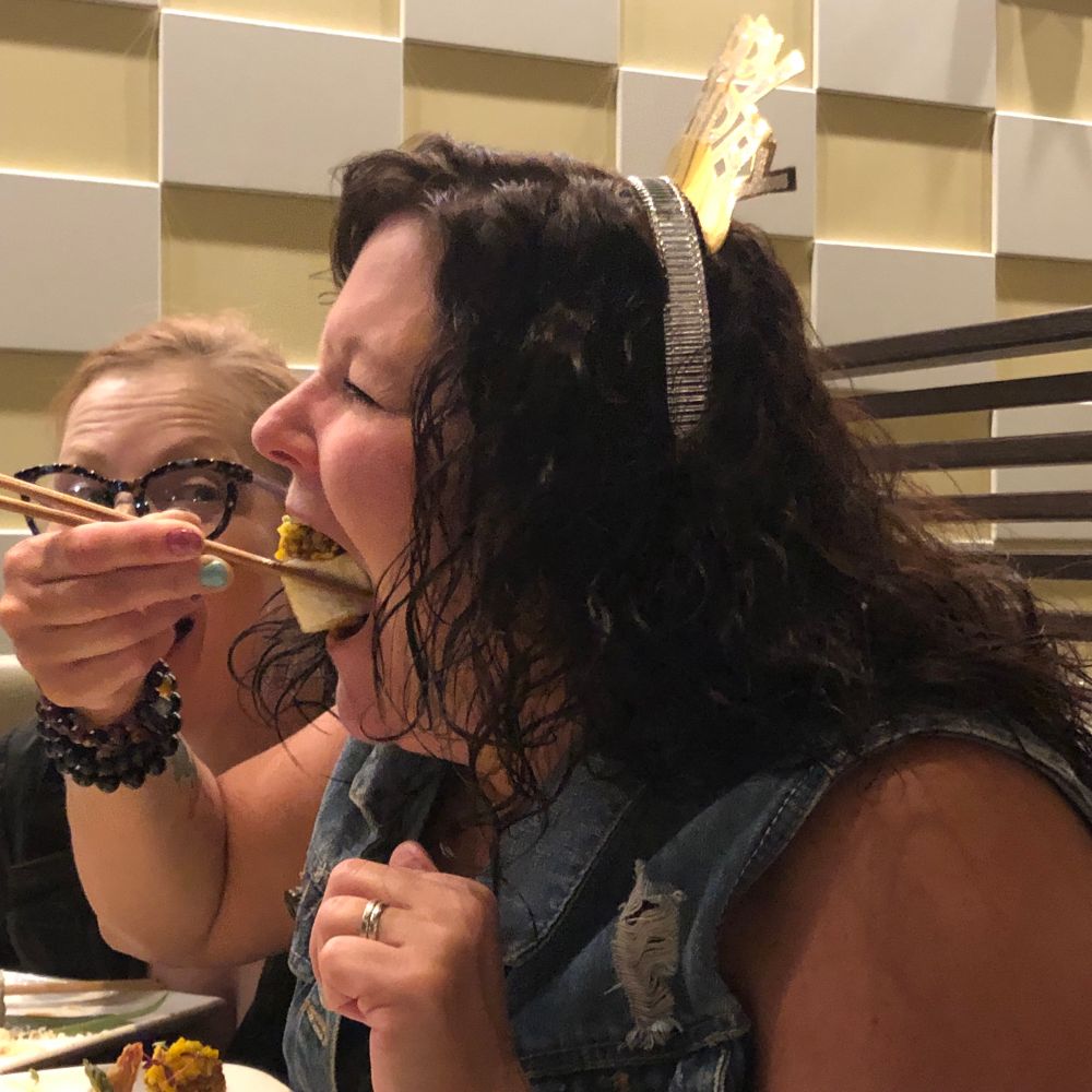 Restaurant Scene: Woman, mouth wide open, placing huge roll in her mouth while an astonished friend looks on. 