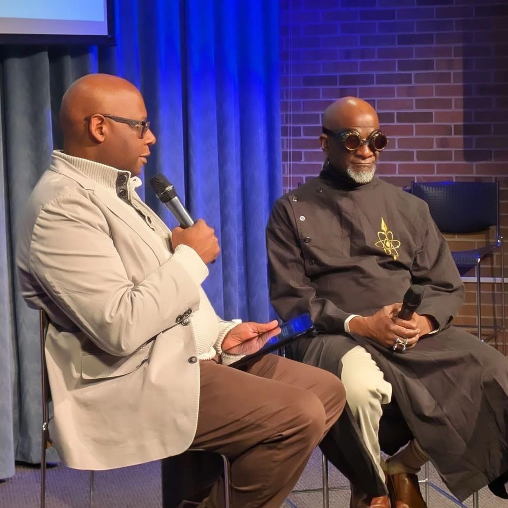 Julian Chambliss seated next to Abdul Qadim Haqq. Both men are looking at each other on a stage and holding microphones. 