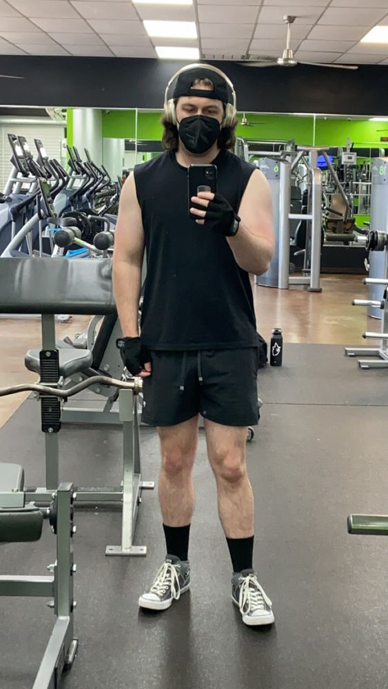 Guy standing, taking selfie in gym mirror. He’s wearing a black sleeveless shirt and black shorts