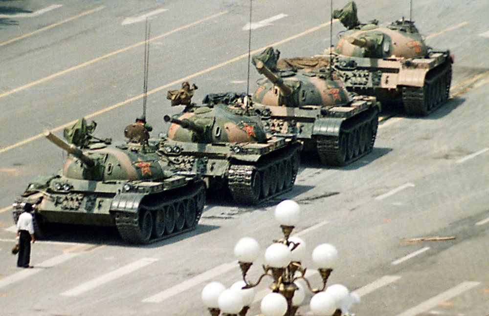 A photo of a man in black pants and white shirt, carrying a grocery bag in his left hand, standing alone in front of a column of four tanks, stopping their progress down an avenue.  A powerful symbol of resistance to the forces moving to Tiananmen Square in Beijing.