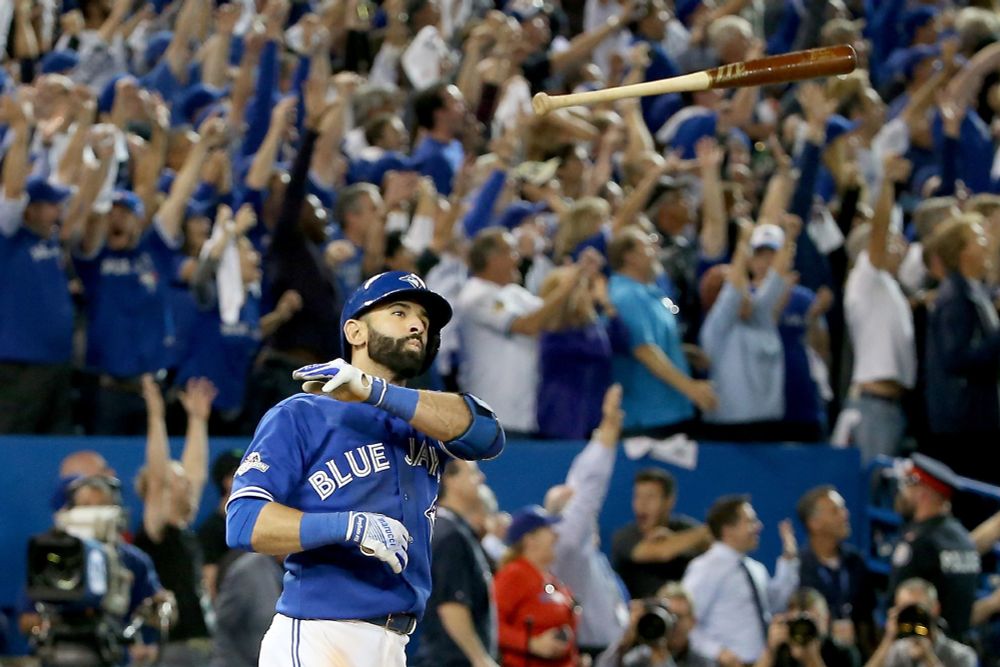 Shot of Jose Bautista, a Toronto Blue Jays outfielder clad in a blue jersey and white pants, tossing his bat high in the air after hitting an historic home run in the 7th inning of Game 5 of the ALDS against the Texas Rangers on October 14, 2015.  The home Rogers Stadium fans behind him are jubilant, standing with arms raised as they watch the flight of the ball on its way into the left field stands.  Joyful scene.