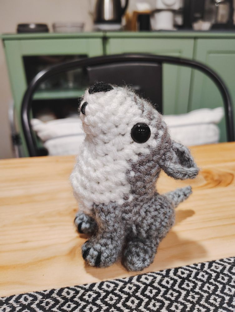 The left side of a small crocheted wolf on a wooden table with a green sideboard in the background. The wolf is gray with a white stomach and black safety eyes and is sitting on its haunches with its nose pointed in the air and ears back as if howling.