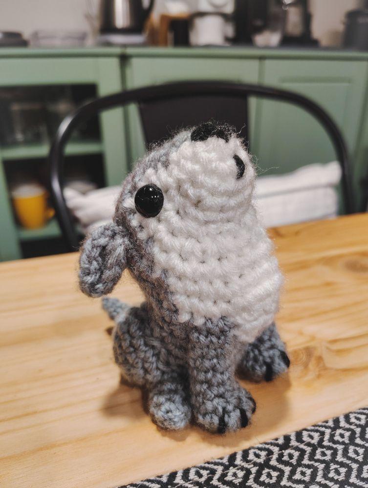 The right side of a small crocheted wolf on a wooden table with a green sideboard in the background. The wolf is gray with a white stomach and black safety eyes and is sitting on its haunches with its nose pointed in the air and ears back as if howling.