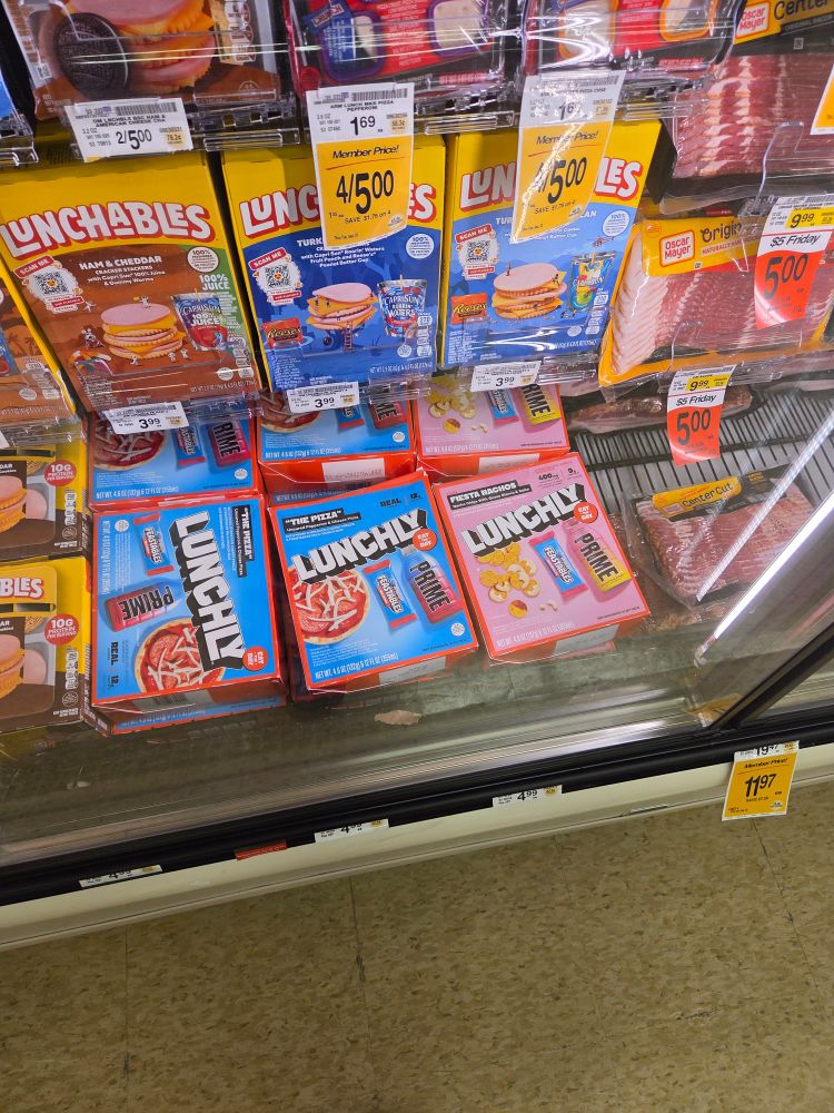 A grocery store's refrigerated display, mostly containing Lunchables boxes, which surround a pitiful 3 rows of boxes of its dollar store knockoff shat out by a trio of shithead "influencers", Lunchly. The Lunchly display is completely full, most likely because its a bad product that even the children who make up the influencers' fanbases don't want.