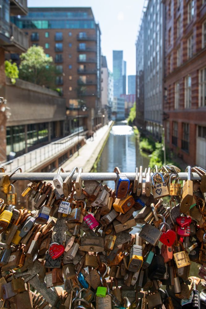 A close-up of dozens of love padlocks attached to a metal railing on a bridge overlooking a canal in Manchester’s city centre. In the background, modern and red-brick buildings line the waterway, with high-rise towers visible in the distance under a bright blue sky.