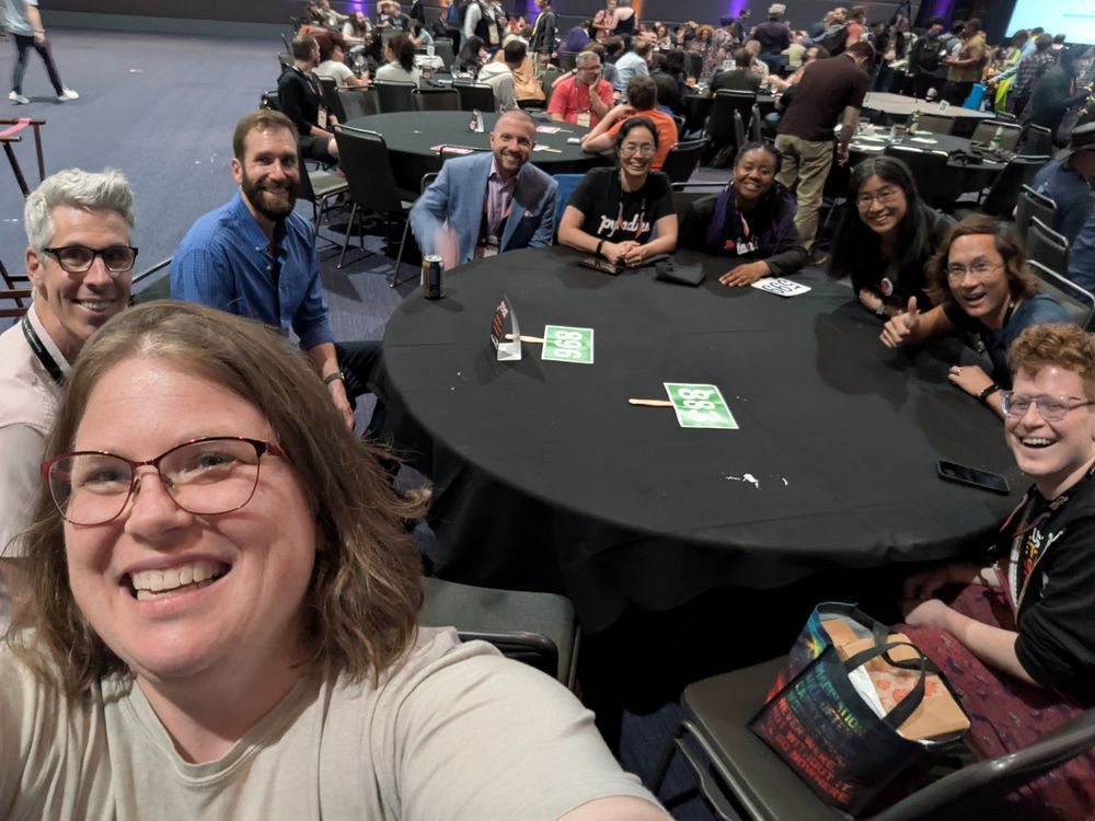 A group of smiling people gathered around a round, black dinner table.