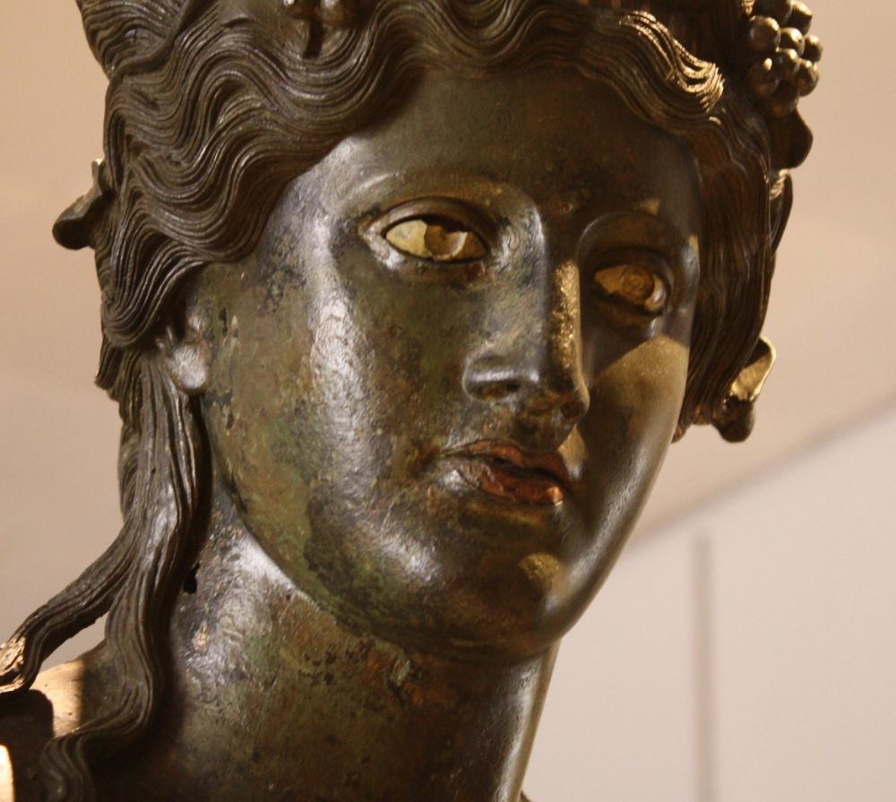 A bronze statue of the god of wine Dionysos (Greek name) / Bacchus (Roman name), early 2nd century CE. His head is crowned with vine leaves and fruit. The eyes are from limestone, the pupils would have probably been in coloured glass paste and the lips are in copper. The statue was made using the lost-wax technique. (Palazzo Massimo, Rome).