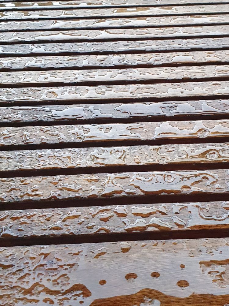 Ein Gartentisch aus Holz, der einem Sommerregen ausgesetzt war. Die kleinen Pfützen bilden unregelmäßige Formen und schimmern. 