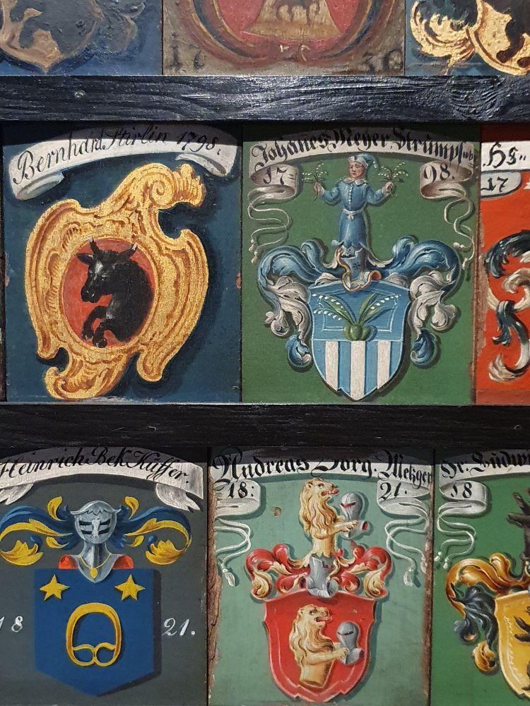 Jeder neue Zünfter hatte ein bemaltes Holzschildchen mit Familienwappen, Vor- und Nachnamen und Jahr der Aufnahme. Diese wurden neben- und übereinander in einem Gestell eingerückt und hingen quasi als Verzeichnis in den Zunftstuben. Es sind 9 Schilder zu sehen. Da sie schräg versetzt sind, zeigt der Ausschnitt nur 4 Wappen vollständig. Alle zeigen weiße Bänder mit dem jeweiligen Familiennamen. Zu sehen ist ein schwarzer Stier auf rotem Grund, daneben ein blaues Wappen mit Maiglöckchen(?), darunter: eine Brezel mit 2 Sternen, und daneben ein Löwe, der einen Ritterhelm in den Pranken hält. 