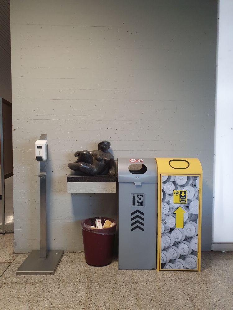 Eine dem/der BetrachterIn zugewandte sitzende Statue aus schwarzem Stein, positioniert auf einem eigens dafür konzipierten Betonvorsprung. Rechts daneben, direkt dran geschoben, 2 Container für PET-Flaschen und Plastik, links daneben ein Desinfektionsspender aus Metall, darunter ein Mülleimer. 