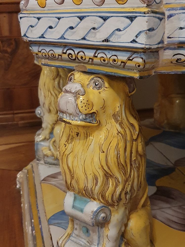 Hier sieht man einen kleinen gelben Löwen, der sich mit seiner Zunge über die Schnauze schleckt. Er trägt mit weiteren Löwen einen prachtvollen Fayencen-Ofen auf dem Kopf, der in der Regel alle Aufmerksamkeit auf sich zieht, so dass man die kleinen Löwen eher übersieht. Der Perspektivenwechsel besteht also im Fokus auf das, was man eher übersieht.