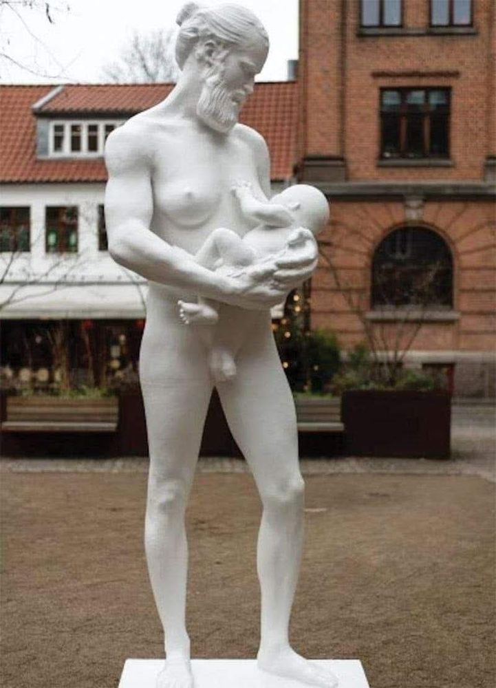 Auf dem Bild ist eine weiße Statue zu sehen, die eine nackte, bärtige Person mit zurückgebundenem Haar zeigt. Die Figur hält ein ebenfalls nacktes Baby im Arm, das an der Brust der erwachsenen Figur zu saugen scheint. Im Hintergrund ist ein Gebäude zu sehen das teils aus rotbrauenen Backsteinen besteht und eine weiß verputzte Wand mit Fenstern.