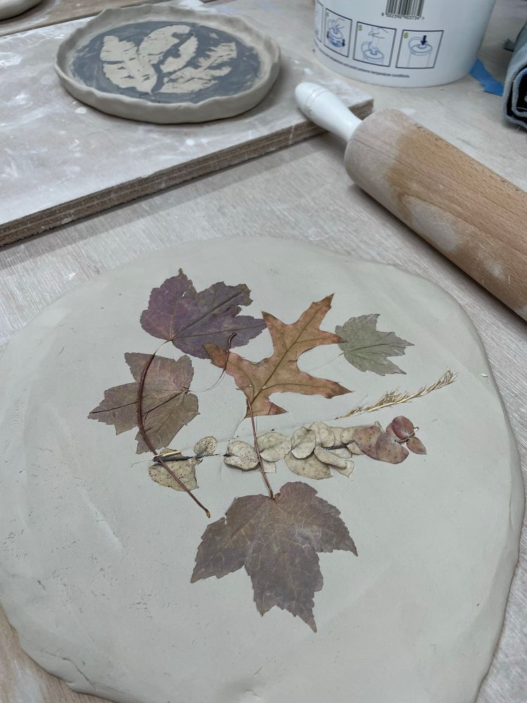 A clay slab with leaves rolled into it. A rolling pin, and a finished imprinted freeform clay plate can be seen in the background. 
