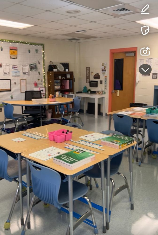 A classroom. There are groups of four desks and a teacher table in the background. 