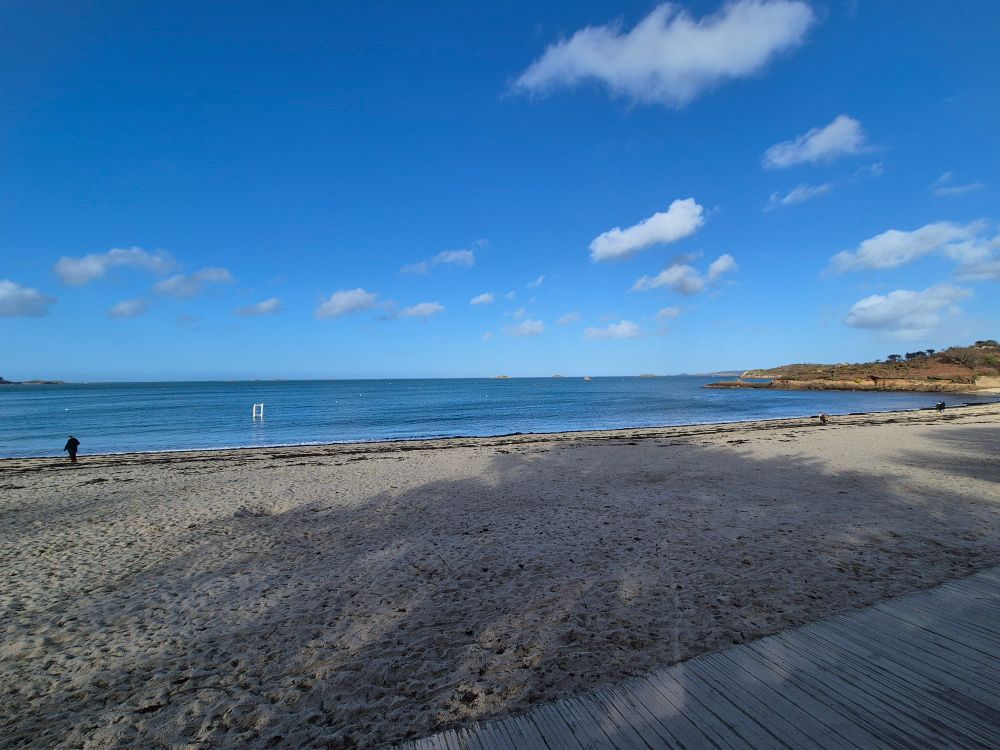 Photo de plage avec ciel bleu. On voit l'ombre d'un arbre sur le sable. Il y a quelques rares nuages blancs dans le ciel. Au premier plan, on voit quelques planches en bois d'une promenade longeant la plage