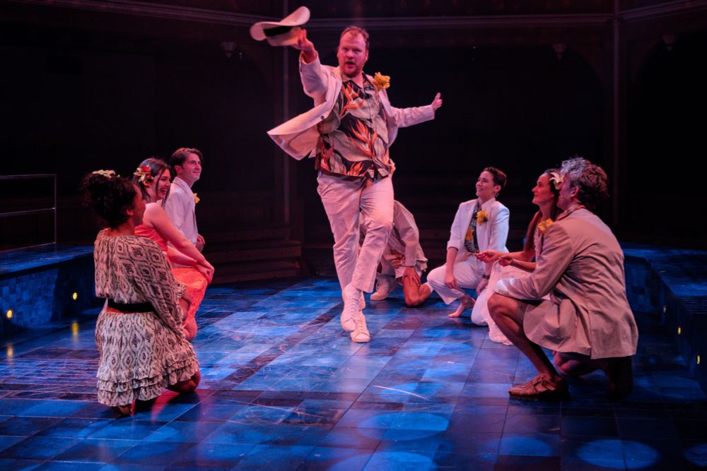 A man twirling a cowboy hat while other people crouch on the floor looking at him. 