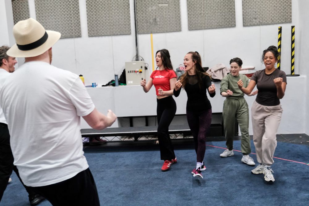 Four women singing and dancing as a group of men look on. 